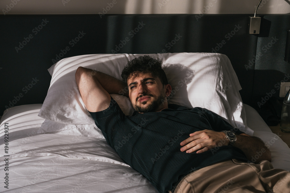 Fototapeta premium Young man lying in bed relaxing during morning sunlight, contemplating and thinking about his life goals. Lifestyle and wellness concept