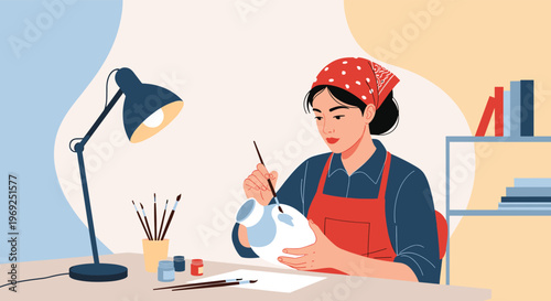 Artistic woman wearing a red headscarf and apron carefully painting a white ceramic vase on her studio desk.