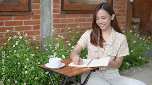A woman is sitting at a table with a notebook and a cup of coffee. She is writing in her notebook and smiling. The scene suggests a relaxed and comfortable atmosphere