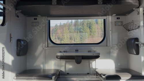 Inside a train compartment, two seats face each other with a small table in between. A window shows trees in the background. Sunlight enters the space, creating a bright atmosphere.