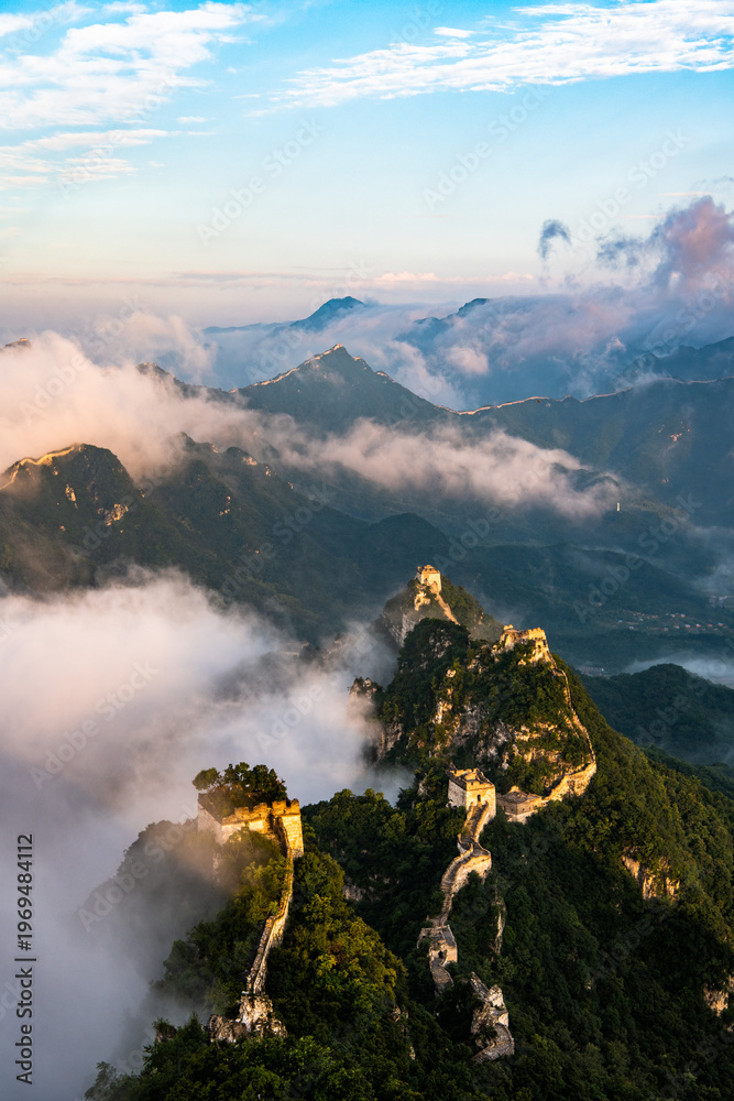 Fototapeta premium Aerial view of the Jiankou section of the Great Wall of China emerging through clouds at sunrise. The ancient wall winds along steep mountain ridges, partially covered by drifting mist