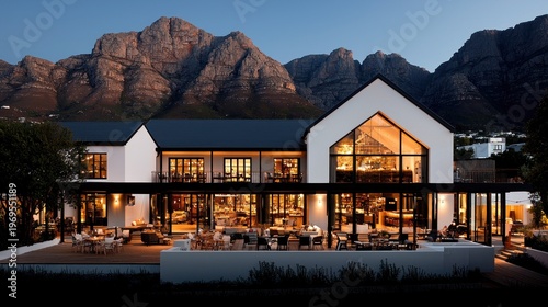 Elegant two-story building with a white facade and dark roof, illuminated interiors, and outdoor dining, set against majestic mountains