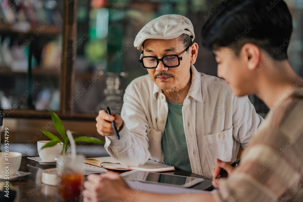 Fototapeta premium Asian father teaches Gen Z son cafe startup accounting at small family business table. SME planning, expense tracking, digital finance learning in cozy coffee shop environment with tablet and notes.