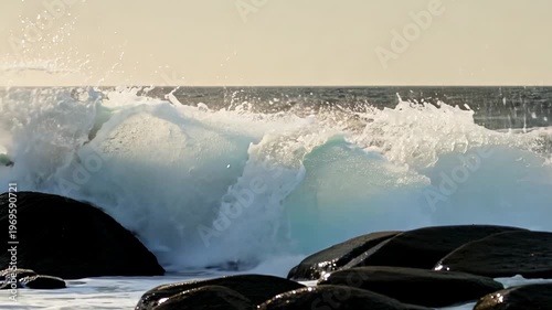 Ocean waves crash against rocky shore, showcasing the dynamic movement of water and foam in a coastal landscape during sunset with soft lighting