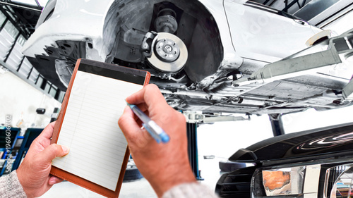 Car Inspection Checklist, A mechanic writes on a blank notepad while performing a vehicle inspection on a car raised on a hydraulic lift in a modern auto garage.