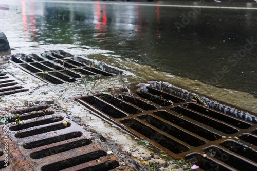 Rainwater runoff moving into a metal sewer grate beside a sidewalk, symbolizing drainage and city maintenance.
