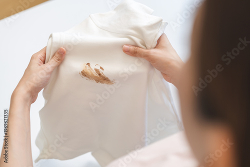 Fotografie Close up hand of housewife, maid woman holding white t-shirt, showing making cloth stain, spot dirty or smudge on clothes, dirt stains for cleaning before washing, making household working at home
