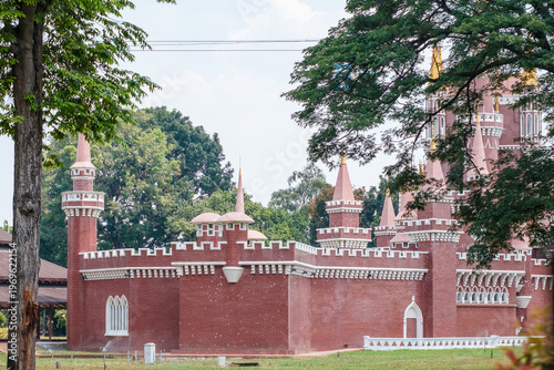 Red brick castle with pointed turrets and white trim. Green trees frame the structure on either side. Bright sky above with light cloud cover. Castle stands on grassy grounds with low fence