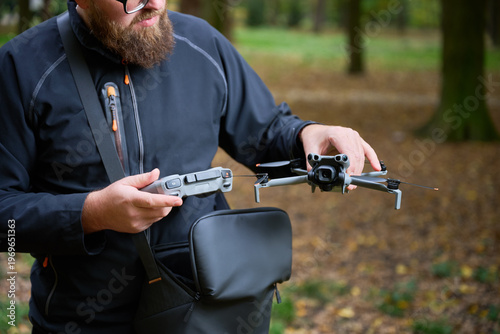 A man with a beard stands in a peaceful park, getting ready to launch his drone. The colorful autumn leaves surround him, creating a tranquil atmosphere.