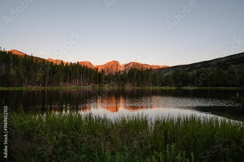 Sprague Lake reflecting sunlit mountains and pine trees at dawn