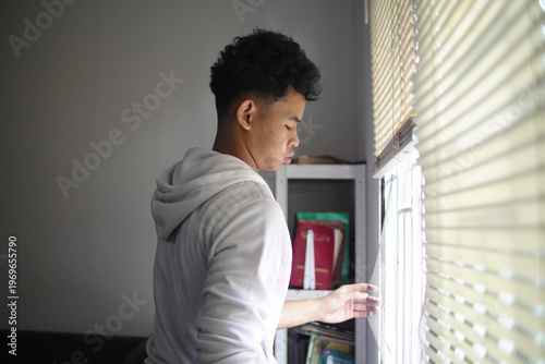 Young Asian man opening or closing window blinds at home, looking out for hope or privacy, morning routine or mental health concept