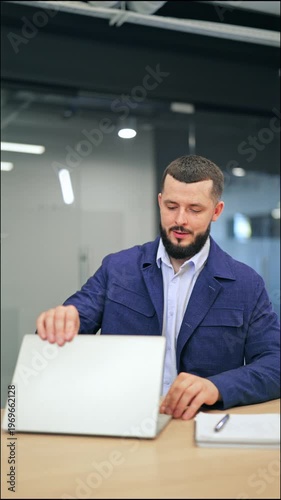 Relieved bearded man in blue jacket closing laptop and stretching hands with satisfaction. Male executive feeling happy and relaxed after finishing tasks or reaching goals in modern office.