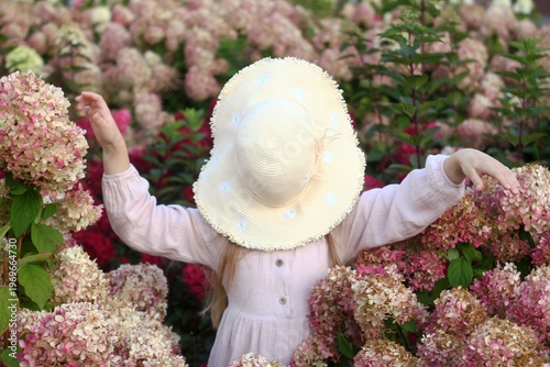 Little girl with charming hat stands in garden with pink and white hydrangeas. The joy of childhood.