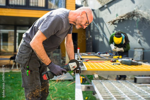Two men are busy working in a backyard. One man cuts metal with a power tool while the other man is focused on a different task nearby. The sun is shining brightly