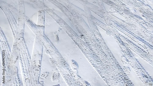 Snow-covered ground with visible tire tracks and footprints creating a dynamic pattern across the white surface, showcasing the interaction between vehicles and pedestrians