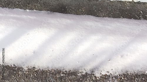 Snow-covered ground with visible shadows cast by nearby objects, showcasing the gradual melting of snow along a gravel pathway in a winter setting