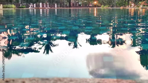 The surface of a swimming pool reflects trees, lights, and a serene sunset sky.