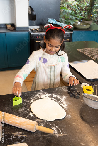 Indian child in striped apron and headband rolling cookie dough with cookie cutters in home kitchen