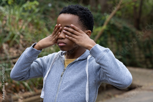 African American child standing on paved park path covering eyes, wearing gray hoodie, yellow tee