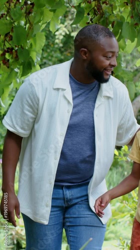Vertical video: Glancing up, father in white shirt in yard guiding son reaching up to fruit in tree