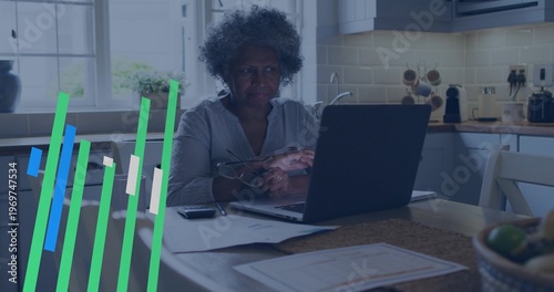 Sitting senior woman wearing grey top with reading glasses, checking papers using laptop in kitchen