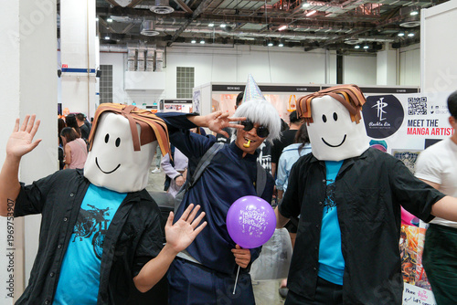 Three people pose with smiling cartoon character masks. One holds a purple balloon. They wave and gesture at a convention event. Bright lights illuminate the indoor space