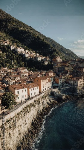 Aerial panoramic view of the historic village of Santa Maria Rezzonico on Lake Como, Italy, featuring traditional colorful houses on rocky cliffs with mountain peaks and blue water.