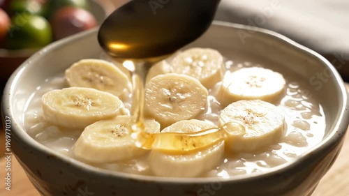 Close-up of sliced bananas being added to a bowl of creamy dessert, followed by a spoon drizzling syrup over the top in a warm kitchen setting
