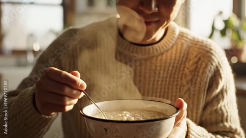 Asian male in cozy sweater enjoys steaming bowl of oatmeal while stirring with a spoon, warm sunlight illuminating the kitchen setting with plants in the background