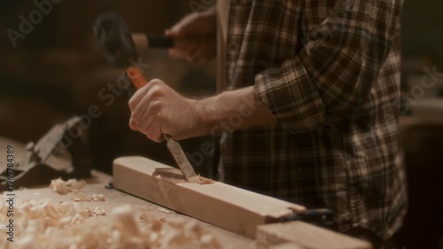 Artisan shaping wooden board with chisel and mallet in workshop, carving material with strikes at workbench in woodworking workshop