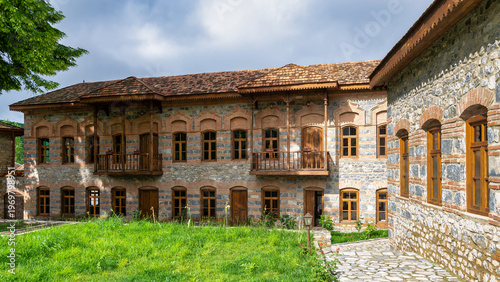 Courtyard of Shaki Khans Mosque Complex in Shaki, Azerbaijan. Historic stone and brick building with wooden balconies, a tiled roof, and green grass under a cloudy sky