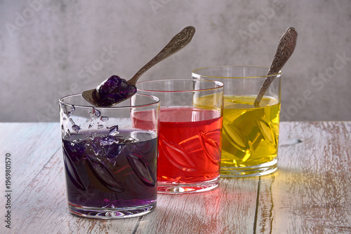 Three clear glasses hold colorful jelly desserts in purple, red, and yellow. A spoon is lifting a piece of purple jelly, attracting attention from the viewers