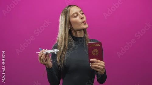 Woman holding passport and model airplane, puckering lips while looking upward against a pink studio wall; wanderlust travel dreams.