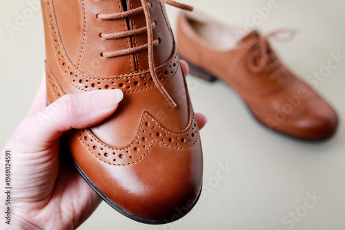 A woman holds a single Oxford shoe in her hand, with the other shoe of the pair visible in the background. The photo metaphorically represents a person appreciating luxury
