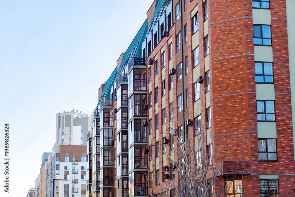 Fototapeta premium Bright red brick building rises beside blue sky. Windows line the facade, some with balconies. Air conditioners hang near every window. Street view shows urban architecture with clean lines