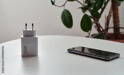 Smartphone and charger on a white table with blurred plant background. Modern mobile technology, charging accessory, and home workspace concept.
