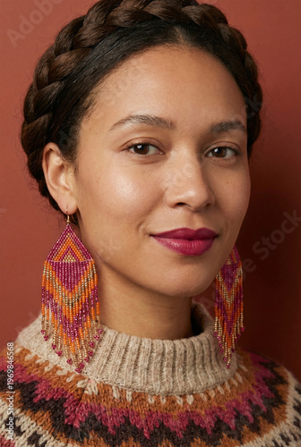 Portrait of a beautiful smiling woman with a crown braid, colorful beaded earrings, and a cozy patterned knit sweater against a warm terracotta background.