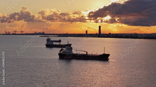 Aerial drone view of multiple oil tankers anchored near an industrial port at sunset with cranes and harbor infrastructure visible on the horizon, Limassol, Cyprus