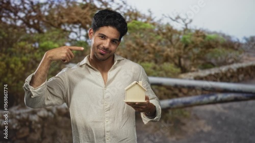 Young man holds miniature wooden house and points finger to chest on a street; homeownership pride aspiration.