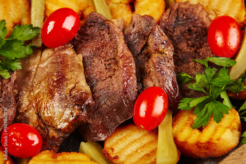 Grilled meat served with roasted fried potatoes, cherry tomatoes, and greens on a hot cast iron skillet with white sauce on a wooden board.