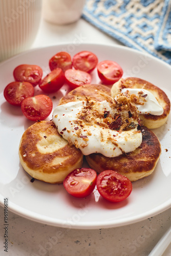 Savory syrniki (cheese pancakes) topped with sour cream, crispy fried onions, and fresh cherry tomatoes, served on a white plate with a blue knit cloth. Perfect for breakfast or brunch.