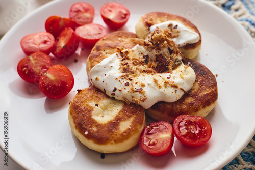 Savory syrniki (cheese pancakes) topped with sour cream, crispy fried onions, and fresh cherry tomatoes, served on a white plate with a blue knit cloth. Perfect for breakfast or brunch.
