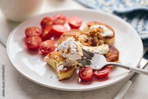 Savory syrniki (cheese pancakes) topped with sour cream, crispy fried onions, and fresh cherry tomatoes, served on a white plate with a blue knit cloth. Perfect for breakfast or brunch.