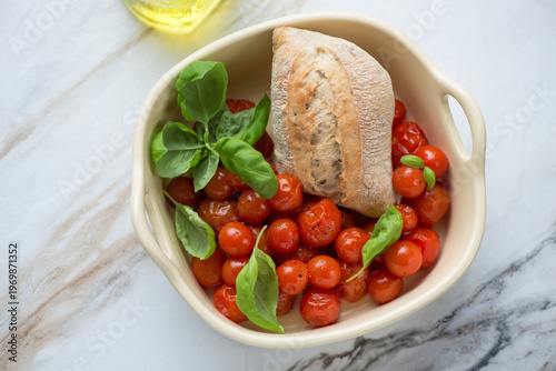 Serving tray with roasted cherry tomatoes, green basil and fresh bread, horizontal shot on a white granite background, high angle view