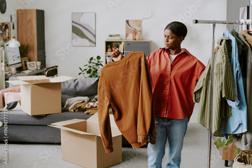 Fotografi African American young adult woman sorting clothing for online resale at home, comparing garments near shipping boxes