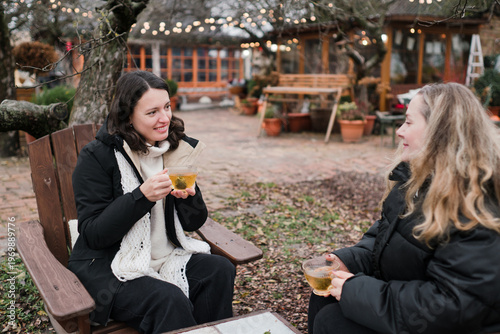 Two pretty caucasian women in their 40s wearing warm clothes spending time outdoors in autumn park cafe. Mature females enjoying personal time, talking, drinking tea, relaxing and breathing fresh air.