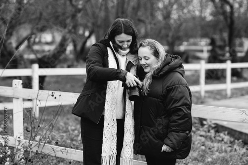 Caucasian woman photographer in her 40s wearing warm clothes taking photos of her friend in autumn park. Relaxed mature females spending time outdoors, enjoying leisure time, walking and posing.