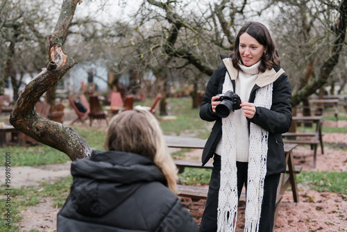 Caucasian woman photographer in her 40s wearing warm clothes taking photos of her friend in autumn park. Relaxed mature females spending time outdoors, enjoying leisure time, walking and posing.