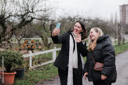 Two pretty caucasian women in their 40s wearing warm clothes walking outdoors in autumn park. Mature female friends enjoying personal time, talking, taking selfies, relaxing and breathing fresh air.