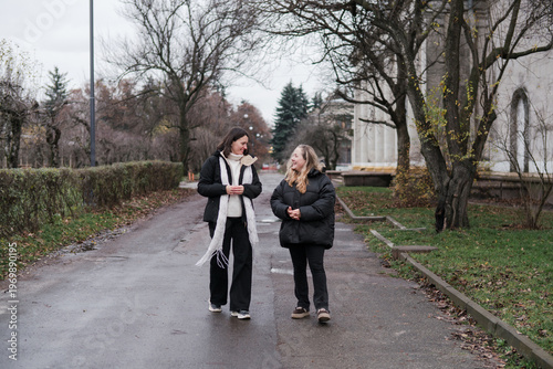 Two pretty caucasian women in their 40s wearing warm clothes walking outdoors in autumn park. Mature female best friends enjoying free leisure time, talking, hugging, relaxing and breathing fresh air.